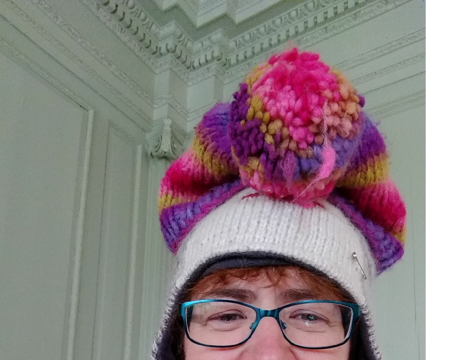 Author wearing a white bobble hat with a pink and purple bobble hat on top; her face is showing from below her blue framed glasses to the top of the hat